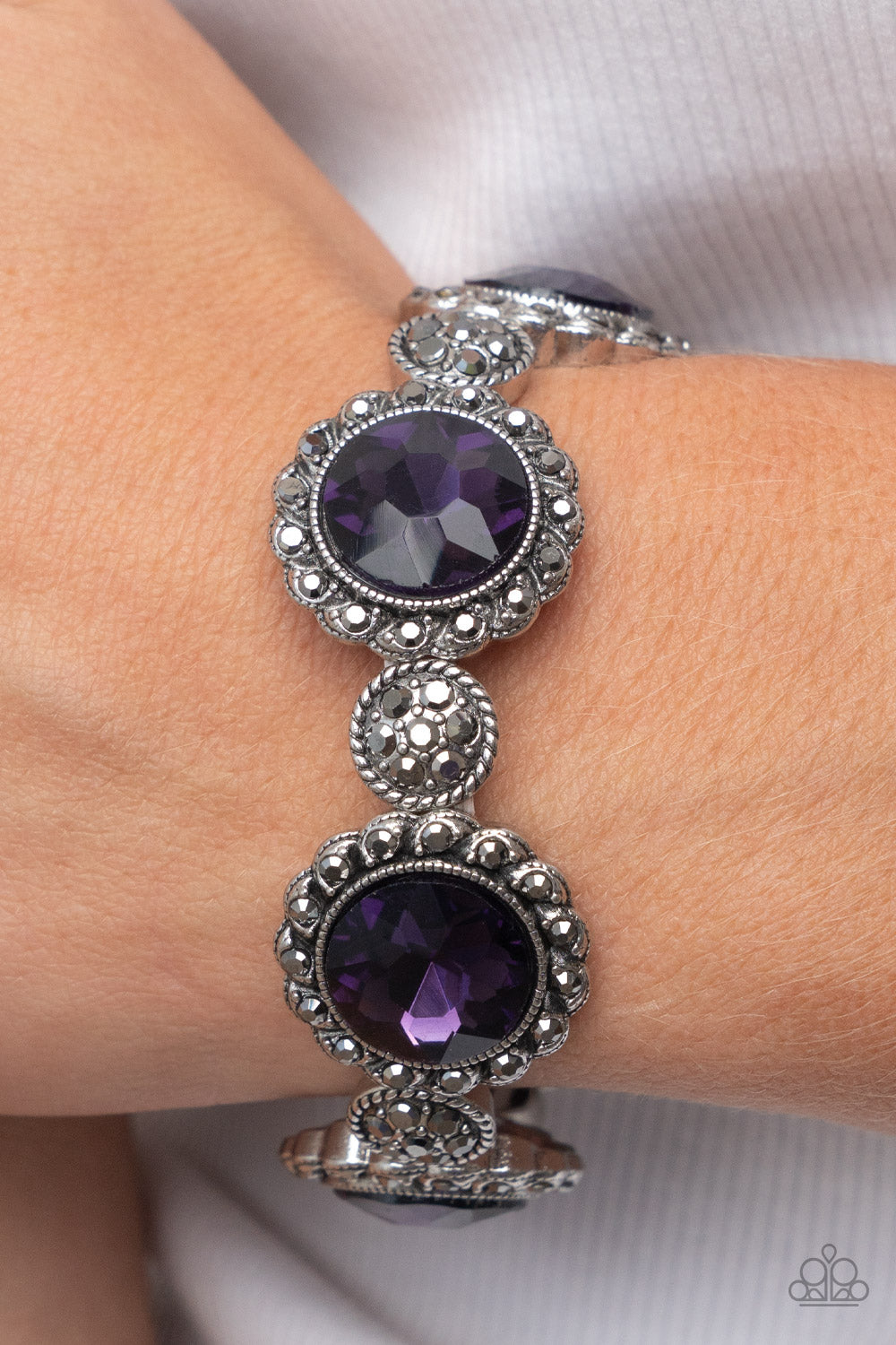 ﻿Palace Property Purple Bracelet - Paparazzi Accessories  An oversized purple rhinestone adorns the center of a hematite rhinestone petaled floral frame. Infused with hematite dotted silver accents, the glitzy floral frames sparkle along stretchy bands around the wrist for a glamorous finish.  Sold as one individual bracelet.