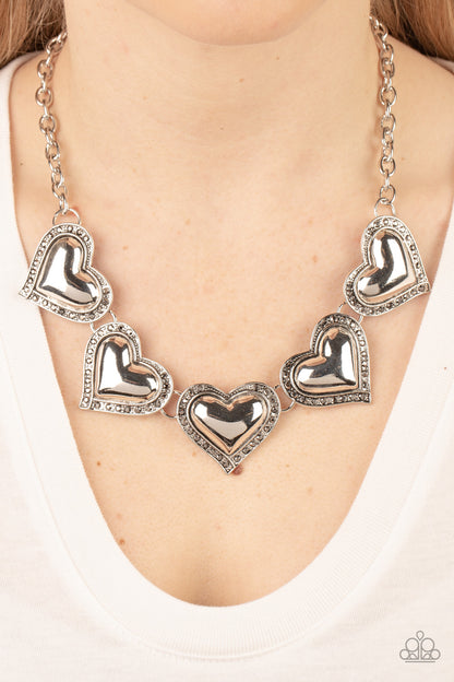 Kindred Hearts Silver Necklace - Paparazzi Accessories  Bordered in glitzy hematite rhinestones, an oversized collection of beveled silver heart frames delicately links below the collar for a dramatically romantic look. Features an adjustable clasp closure.  Sold as one individual necklace. Includes one pair of matching earrings.