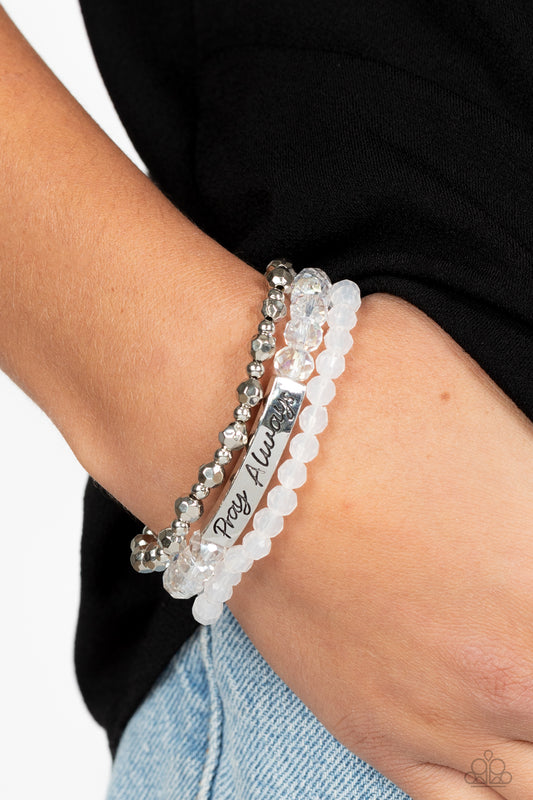 Pray Always White Bracelet - Paparazzi Accessories  Varying in shape and shimmer, faceted silver beads, milky white beads, and glittery crystal-like beads are threaded along elastic stretchy bands, creating colorful layers across the wrist. Featured on the crystal-like band, a curved silver bar features the words "Pray Always" in a curly, stamped font for a soft finish.  Sold as one individual bracelet.